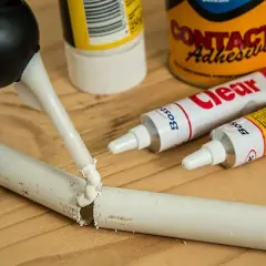 Klej w tubce jest aplikowany na pękniętą białą rurkę. Obok leżą tubki z klejem i puszka "Contact Adhesive". Czy kropelka sklei plastik?