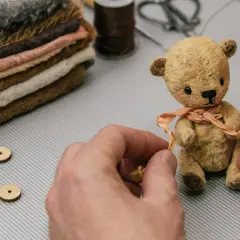 Ręce tworzą uroczego misia, pokazując jak zrobić pluszaka. Stosik futerka i akcesoria czekają na kolejne etapy.