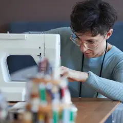 Kobieta w okularach szyje na maszynie, przygotowując lamówkę do obszywania sukien.