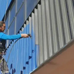 Pracownik na wysokości maluje elewację budynku farbą metaliczną.