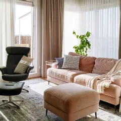 Przytulny salon z kanapą w odcieniu millennial pink, czarnym fotelem i pufą. Jasne zasłony wpuszczają dużo światła.