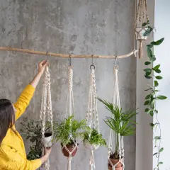 Kobieta w żółtym swetrze wiesza rośliny w makramowych kwietnikach DIY. Dekoracja ścienna z naturalnych materiałów.