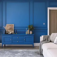 Elegancki salon z boazerią w kolorze royal blue. Na środku niebieska komoda, obok biała kanapa z szarymi poduszkami i pledem.