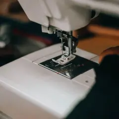 Zbliżenie na białą maszynę do szycia, gdzie igła z pomarańczową nitką przepuszcza ścieg przez materiał.