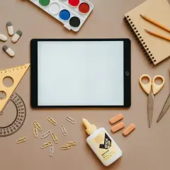 Na stole leżą przybory szkolne: tablet, zeszyt, kredki, nożyczki (jedne z nich to nożyczki dla leworęcznych, czym się różnią? Inaczej wyprofilowane ostrza), linijka, klej i spinacze.