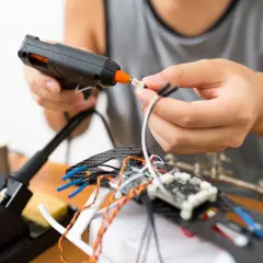Ręce składają elektronikę, używając pistoletu na gorący klej. To szybki sposób, czym skleić metal i inne materiały przy majsterkowaniu.