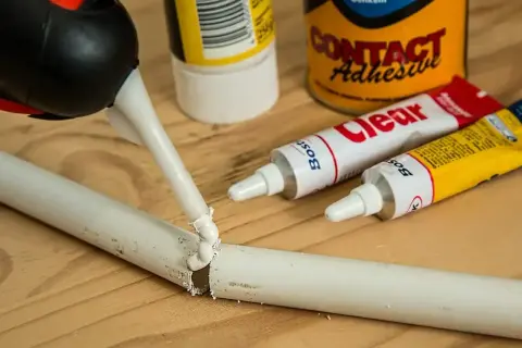 Klej w tubce jest aplikowany na pękniętą białą rurkę. Obok leżą tubki z klejem i puszka "Contact Adhesive". Czy kropelka sklei plastik?