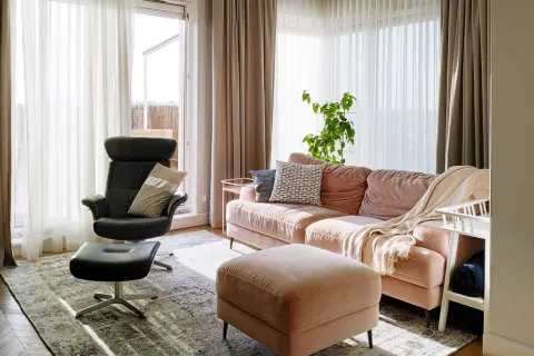 Przytulny salon z kanapą w odcieniu millennial pink, czarnym fotelem i pufą. Jasne zasłony wpuszczają dużo światła.