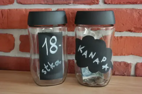 Dwie skarbonki ze słoików. Jedna z napisem "18. stka", druga z napisem "Kanapa" i widocznymi monetami oraz banknotem.