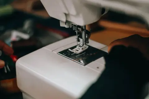 Zbliżenie na białą maszynę do szycia, gdzie igła z pomarańczową nitką przepuszcza ścieg przez materiał.