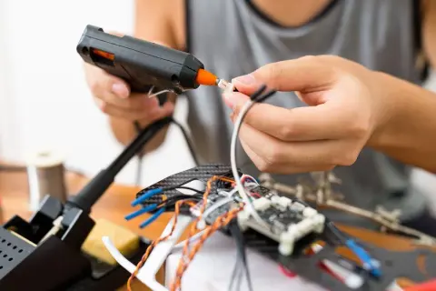 Ręce składają elektronikę, używając pistoletu na gorący klej. To szybki sposób, czym skleić metal i inne materiały przy majsterkowaniu.