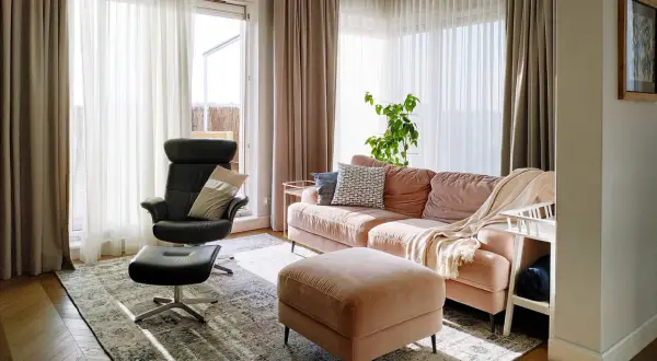 Przytulny salon z kanapą w odcieniu millennial pink, czarnym fotelem i pufą. Jasne zasłony wpuszczają dużo światła.