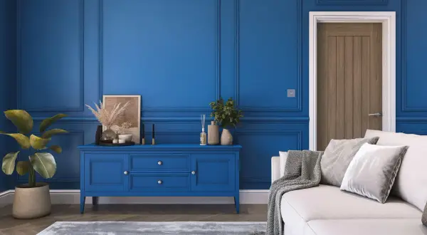 Elegancki salon z boazerią w kolorze royal blue. Na środku niebieska komoda, obok biała kanapa z szarymi poduszkami i pledem.