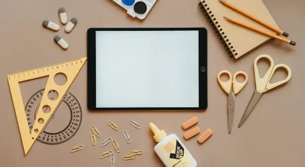Na stole leżą przybory szkolne: tablet, zeszyt, kredki, nożyczki (jedne z nich to nożyczki dla leworęcznych, czym się różnią? Inaczej wyprofilowane ostrza), linijka, klej i spinacze.