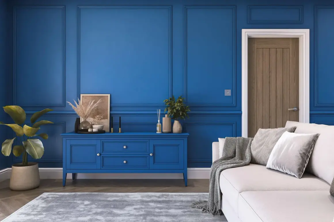 Elegancki salon z boazerią w kolorze royal blue. Na środku niebieska komoda, obok biała kanapa z szarymi poduszkami i pledem.