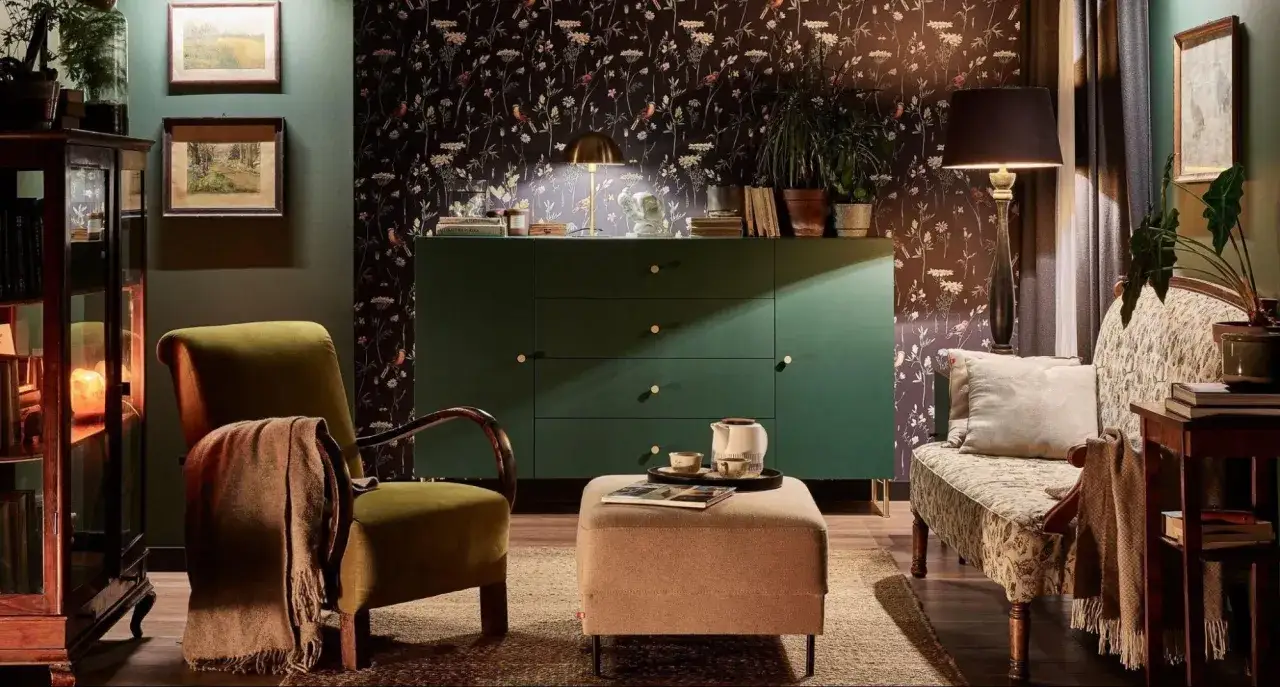 Przytulny salon z zieloną komodą, fotelami i sofą. Na stoliku filiżanki i dzbanek. W tle tapeta z motywem roślinnym, kt&oacute;ra nadaje wnętrzu lekko brudne złoto.