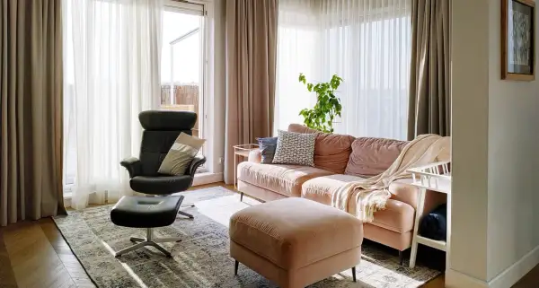 Przytulny salon z kanapą w odcieniu millennial pink, czarnym fotelem i pufą. Jasne zasłony wpuszczają dużo światła.