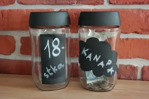 Dwie skarbonki ze słoików. Jedna z napisem "18. stka", druga z napisem "Kanapa" i widocznymi monetami oraz banknotem.