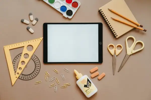 Na stole leżą przybory szkolne: tablet, zeszyt, kredki, nożyczki (jedne z nich to nożyczki dla leworęcznych, czym się różnią? Inaczej wyprofilowane ostrza), linijka, klej i spinacze.
